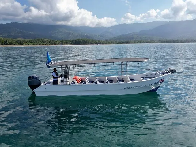 Un pequeño barco turístico blanco flota sobre aguas oceánicas tranquilas cerca de colinas boscosas.